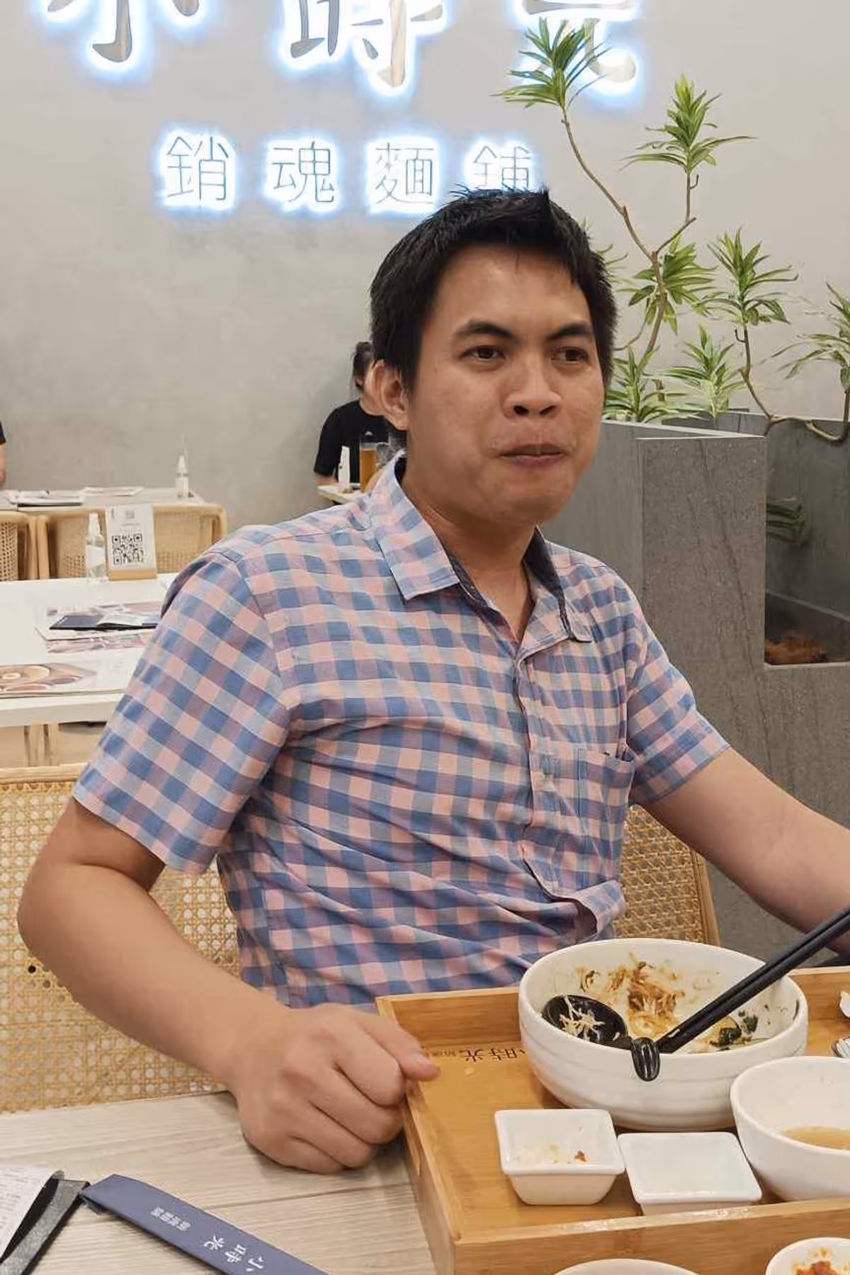 A man in blue and pink flannel sits on a chair with a noodle cup on the table.