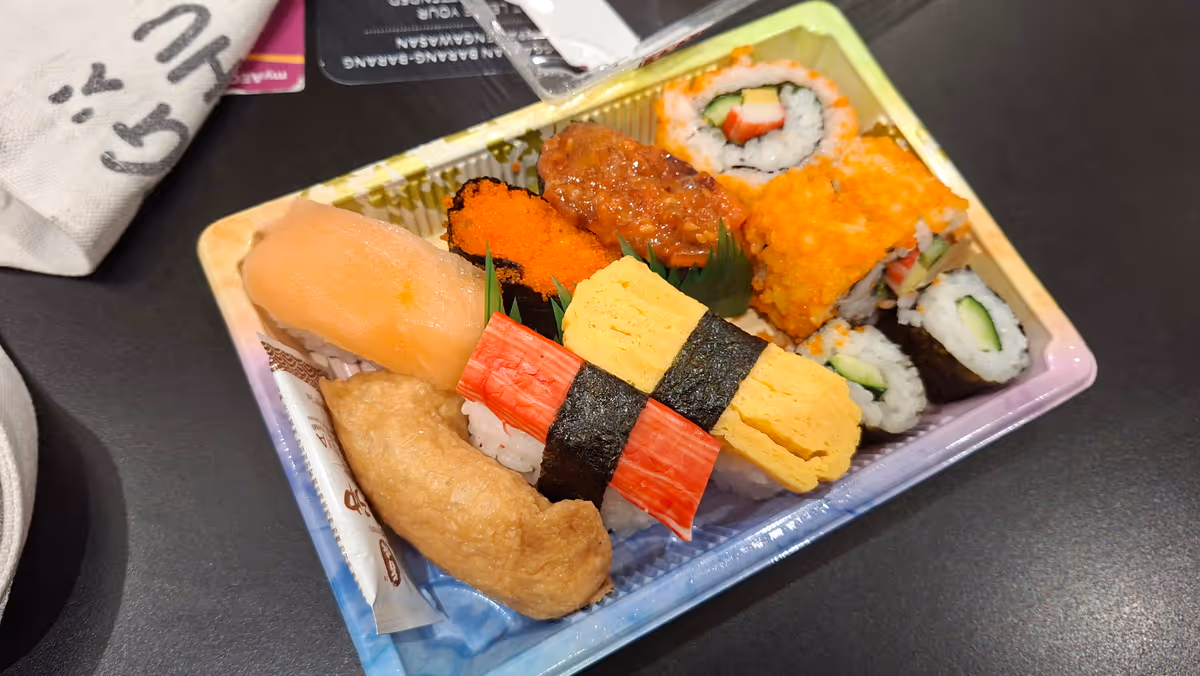 A tray of assorted sushi including salmon nigiri, ebiko sushi, California rolls, inari sushi, crab stick, tamago sushi, kappamaki, and gunkan maki