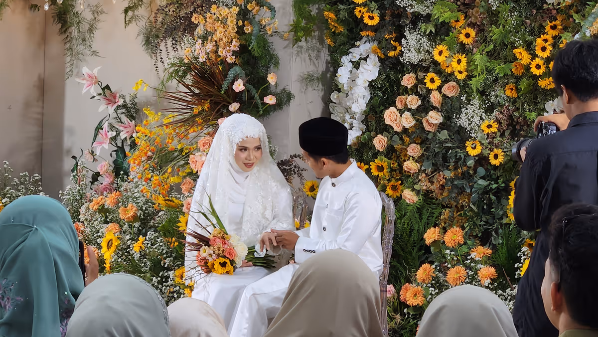 A bride and a groom in white clothes looking at each other. There is a wall of flower in the background.