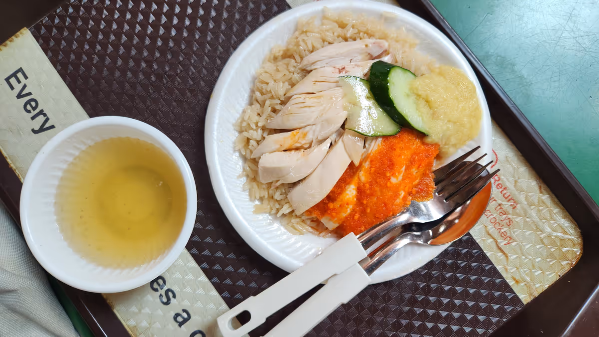 A dish of boil chicken rice and a slice of cucumber, utensils, mashed ginger, red sauce and soup.