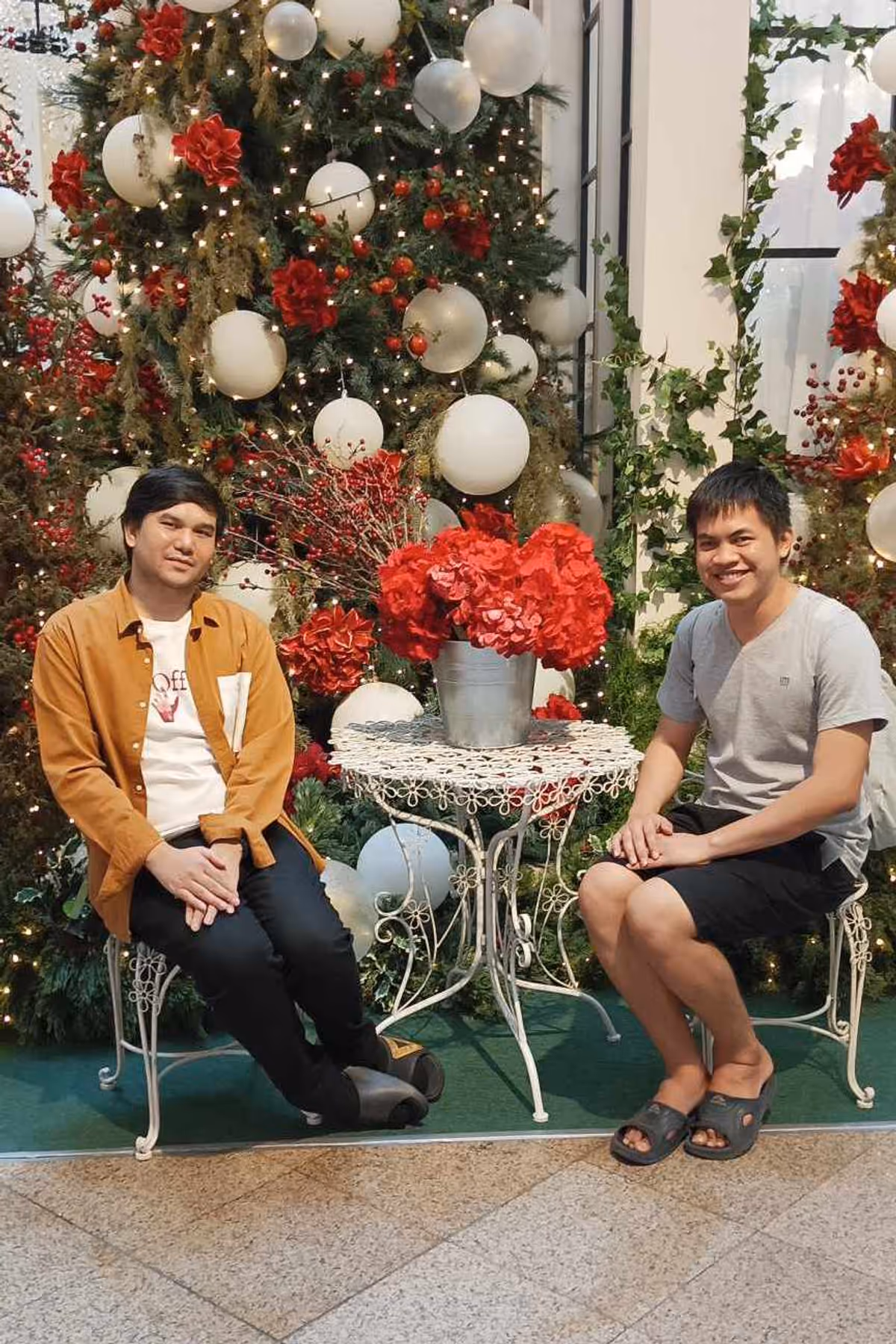 2 people sitting in chairs with a Christmas themed decoration. There is a red flower among them.