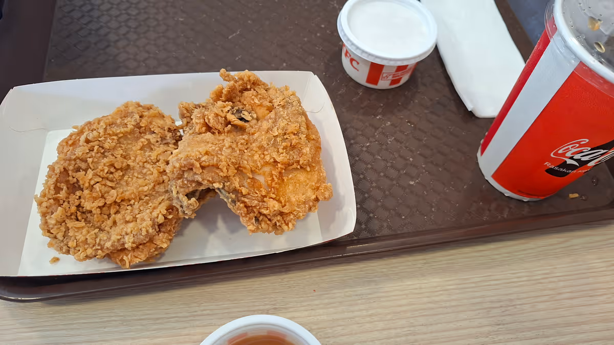 Two pieces of fried chicken in a paper tray, with a lidded paper cup and a small round container on the right side