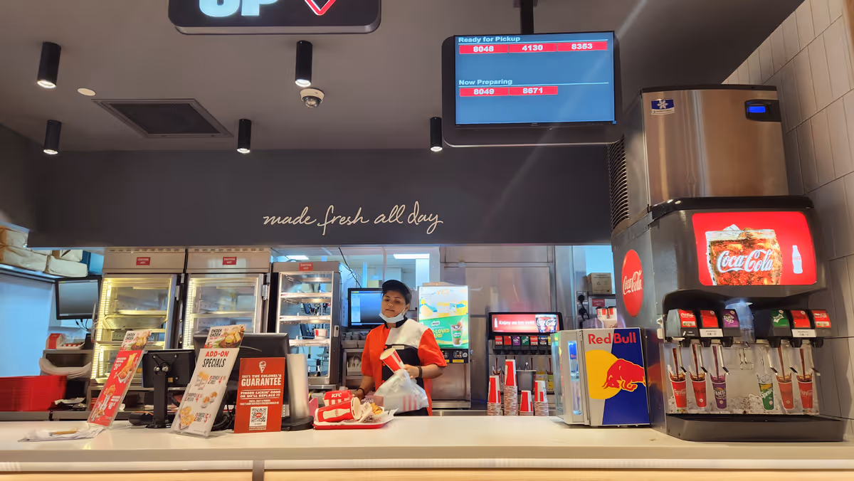 A food stall with a woman at the center and an electronics display. There is a soda fountain on the right.