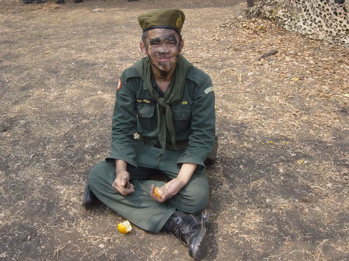 A man in a green military uniform sits on the ground. He holds an orange. His face is covered with camouflage face paint.