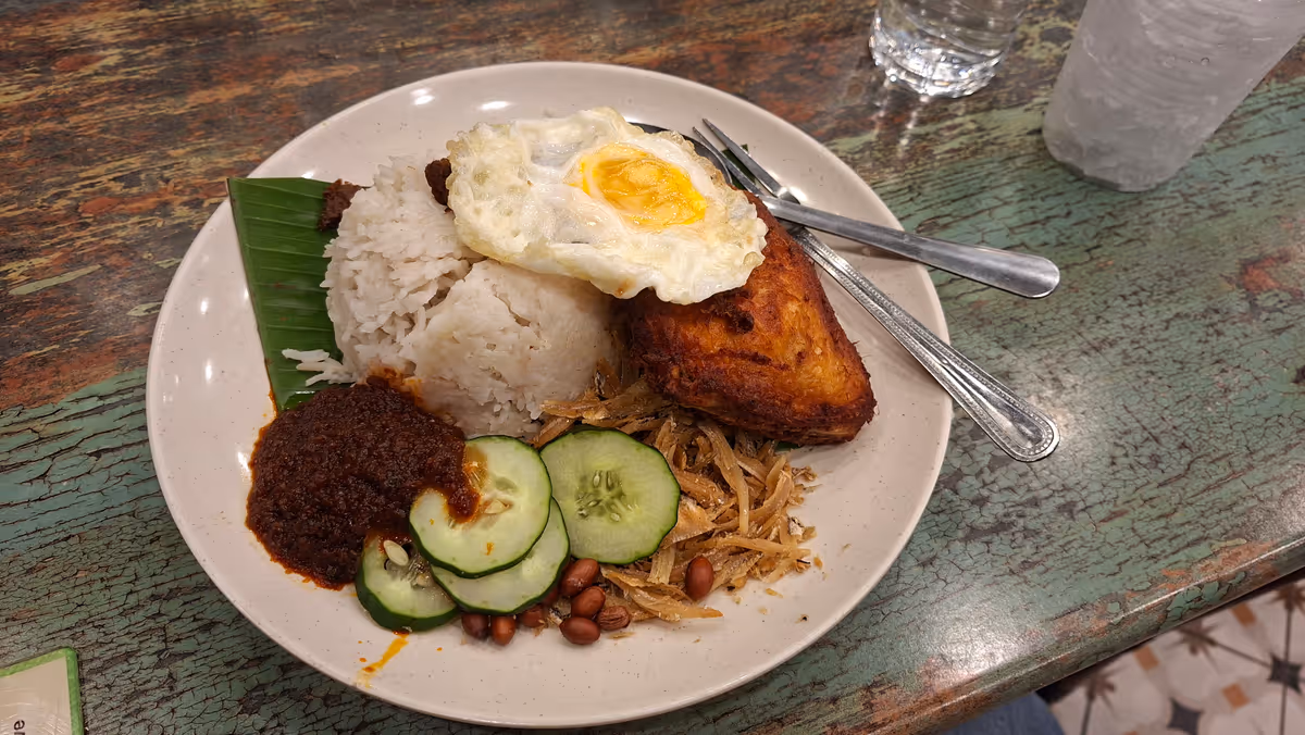 A dish of nasi lemak served on a banana leaf, with chili paste, fried peanuts, dried anchovies, fried egg, cucumber slices, and fried chicken