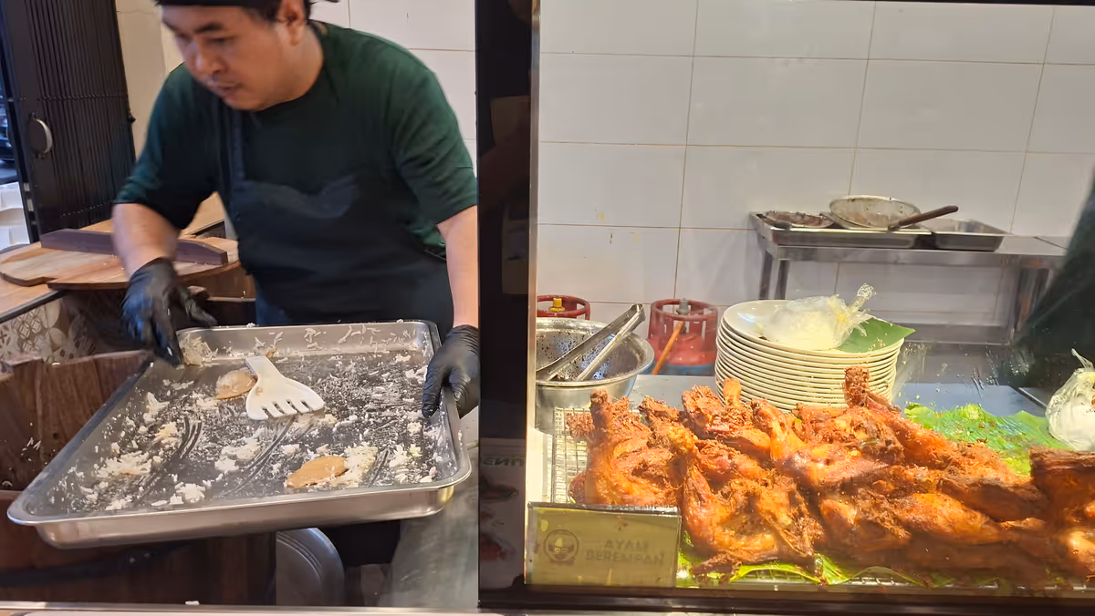A man prepares to cook rice while holding a tray, with a separate tray of fried chicken on the right side behind a transparent partition