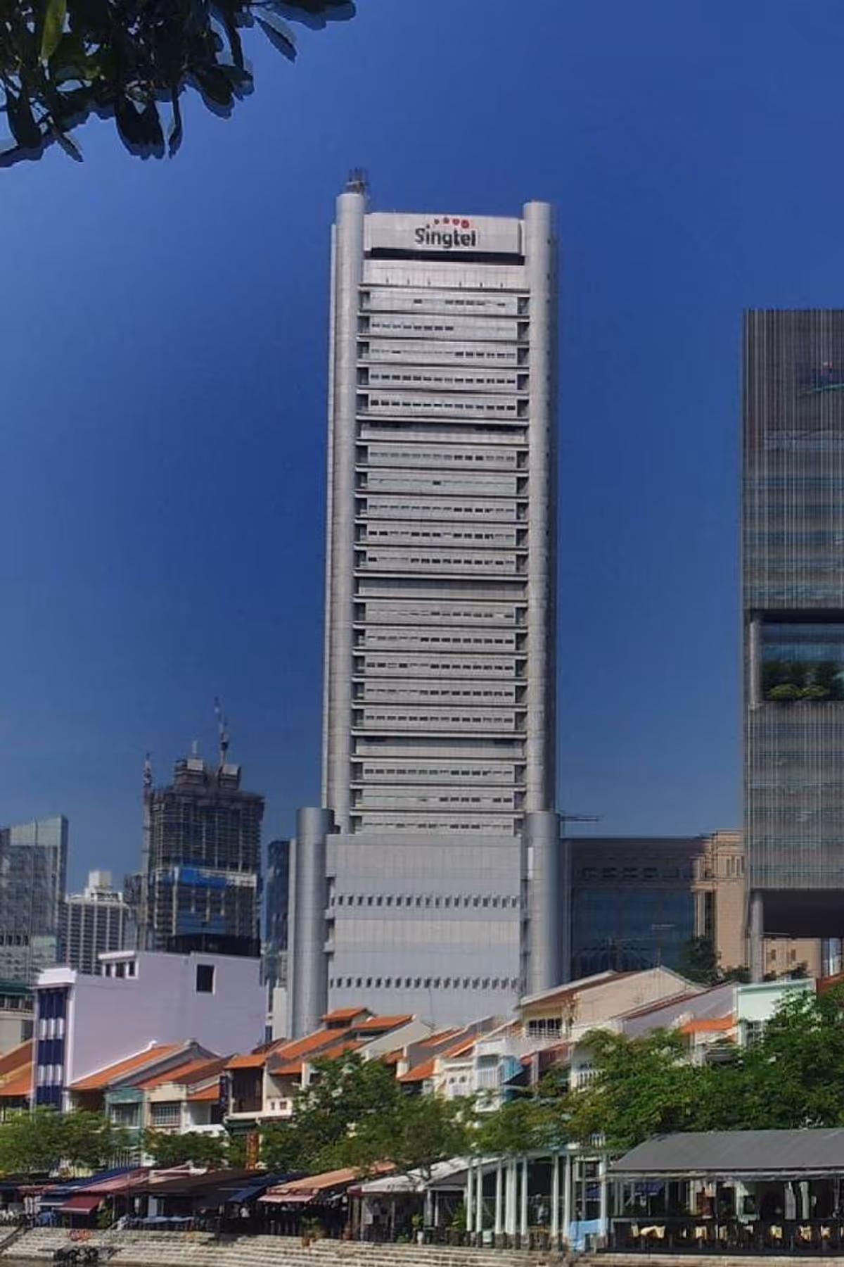 A skyscraper with Singtel with blue sky. There are houses and shop lots in front of the building.