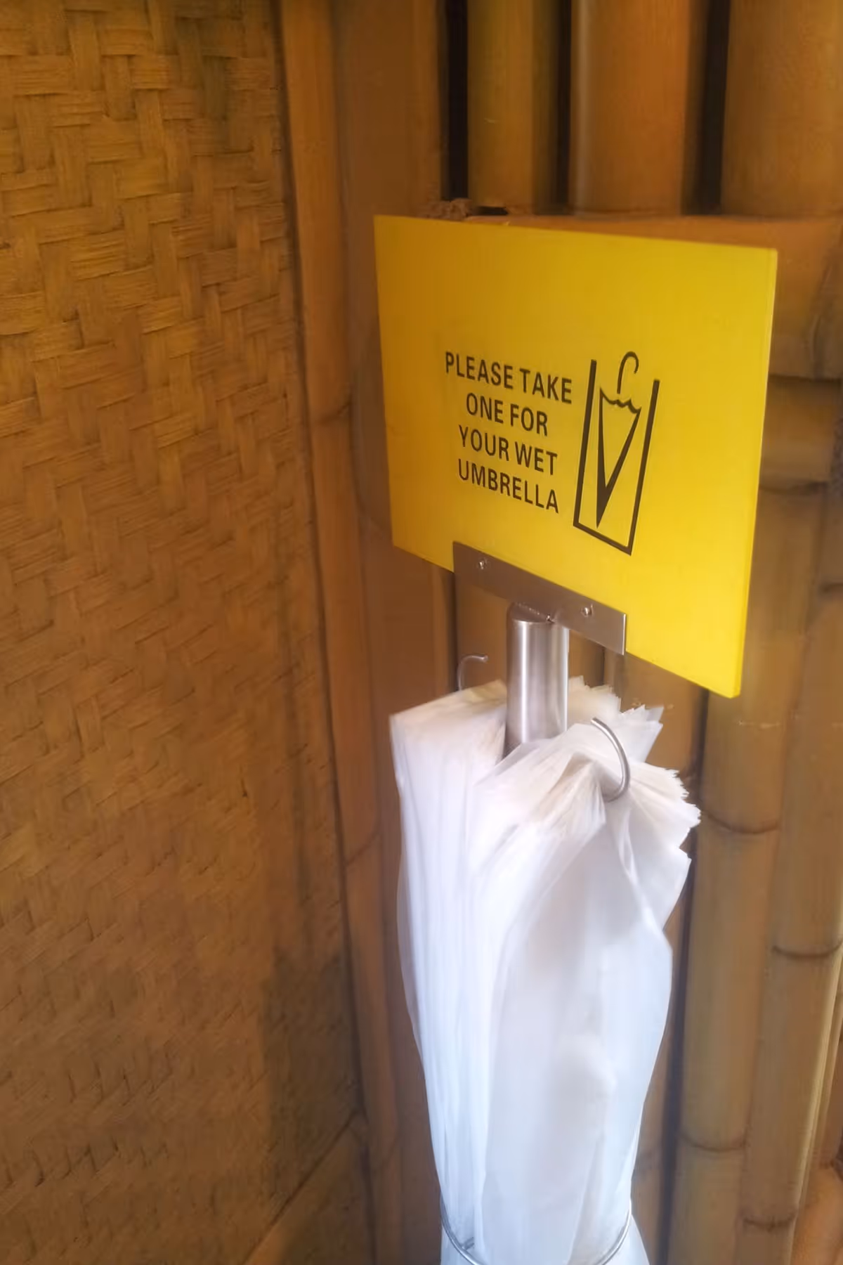 A wet umbrella bag dispenser with a yellow sign board. The wall is made with woven bamboo.
